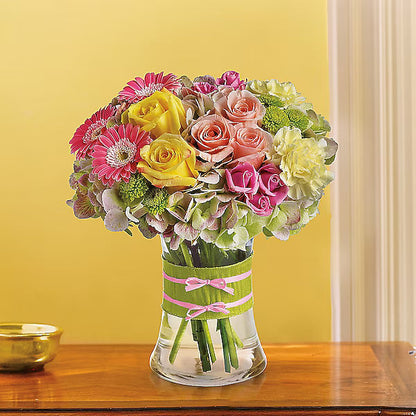 Mixed flowers in clear vase with yellow wall behind.