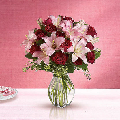 Bouquet of red roses and pink lilies in a clear vase on a pink background