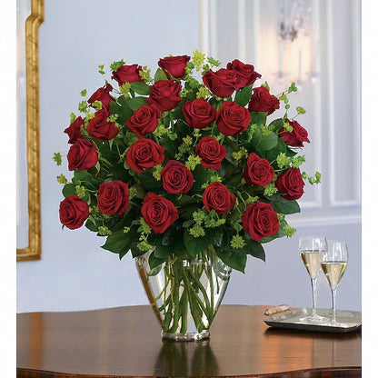 Bouquet of red roses in a clear vase on a table with two wine glasses.